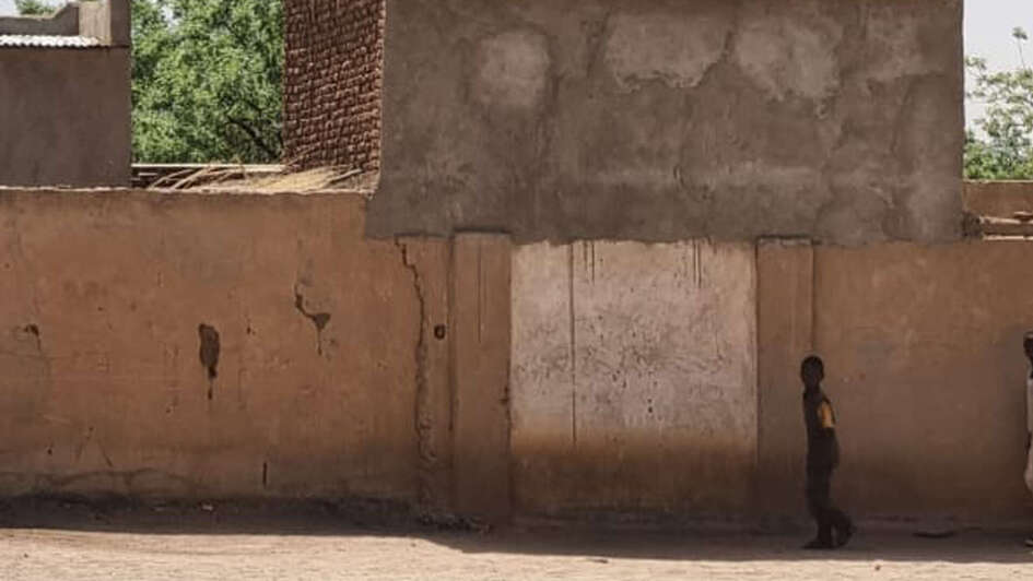 Child standing in front of a house in Sudan after the outbreak of war, smoke rising.