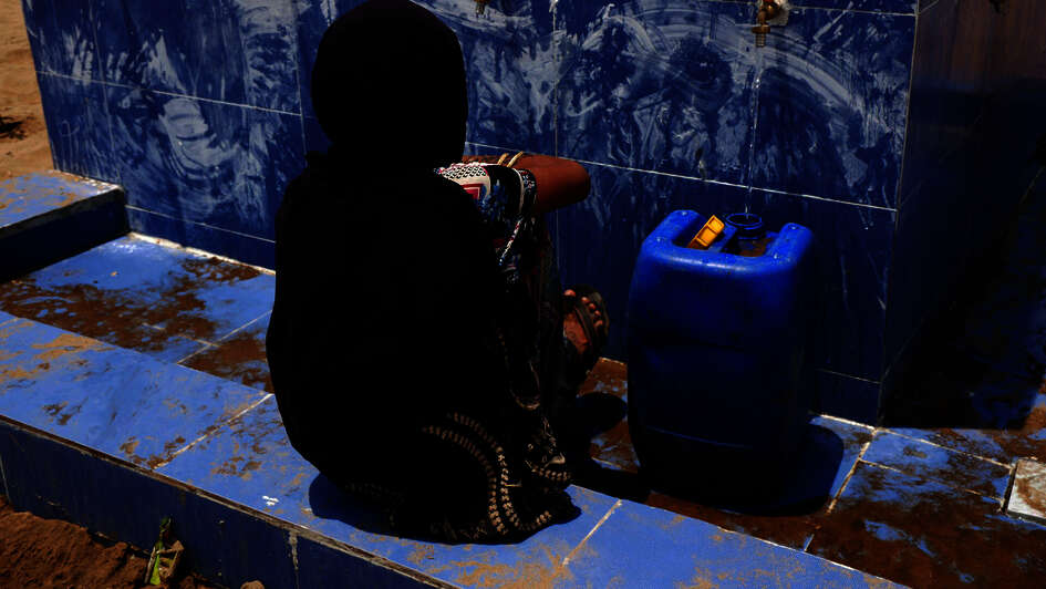 Eine Frau wartet an einer Wasserstelle darauf, dass ihr Kanister mit Trinkwasser voll wird.