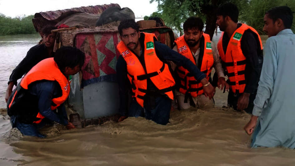 Überschwemmungen in Pakistan. Männer mit Schwimmwesten tragen einen großen Container durch kniehohes Wasser