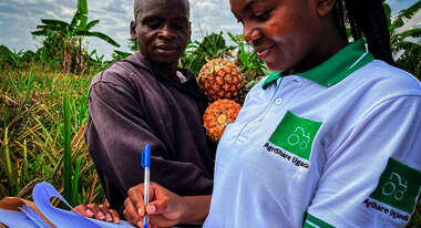 Titelfoto des Jahresbericht 2021. Eine Welthungerhilfe-Mitarbeiterin mit AgriShare-T-Shirt macht Notizen, während ein Mann im Hintergrund zuschaut. Er hält zwei Ananas in den Händen.