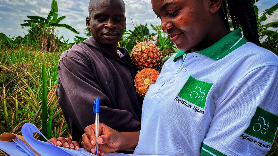 Titelfoto des Jahresbericht 2021. Eine Welthungerhilfe-Mitarbeiterin mit AgriShare-T-Shirt macht Notizen, während ein Mann im Hintergrund zuschaut. Er hält zwei Ananas in den Händen.
