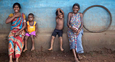 Zwei Frauen und zwei Kinder sitzen an eine Wand gelehnt auf dem Dorfplatz.